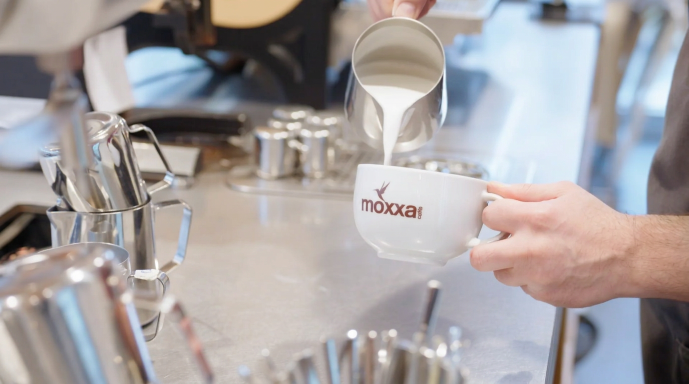 Aufgeschäumter Haferdrink wird aus einem Edelstahlkännchen in eine Kaffeetasse gegossen