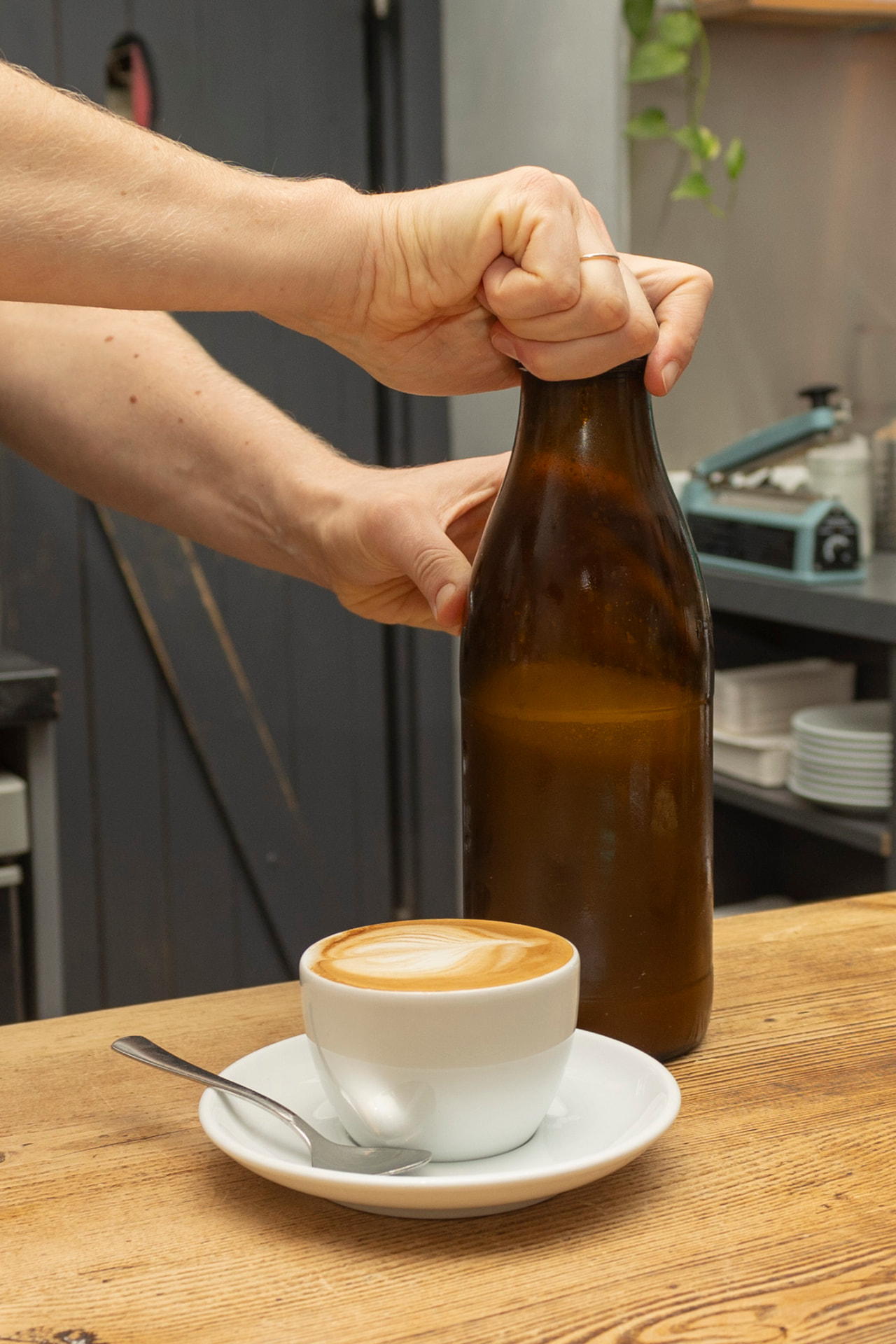 Hand öffnet eine braune Glasflasche neben einer Tasse Haferdrink mit Milchschaum-Muster auf einer Holztheke