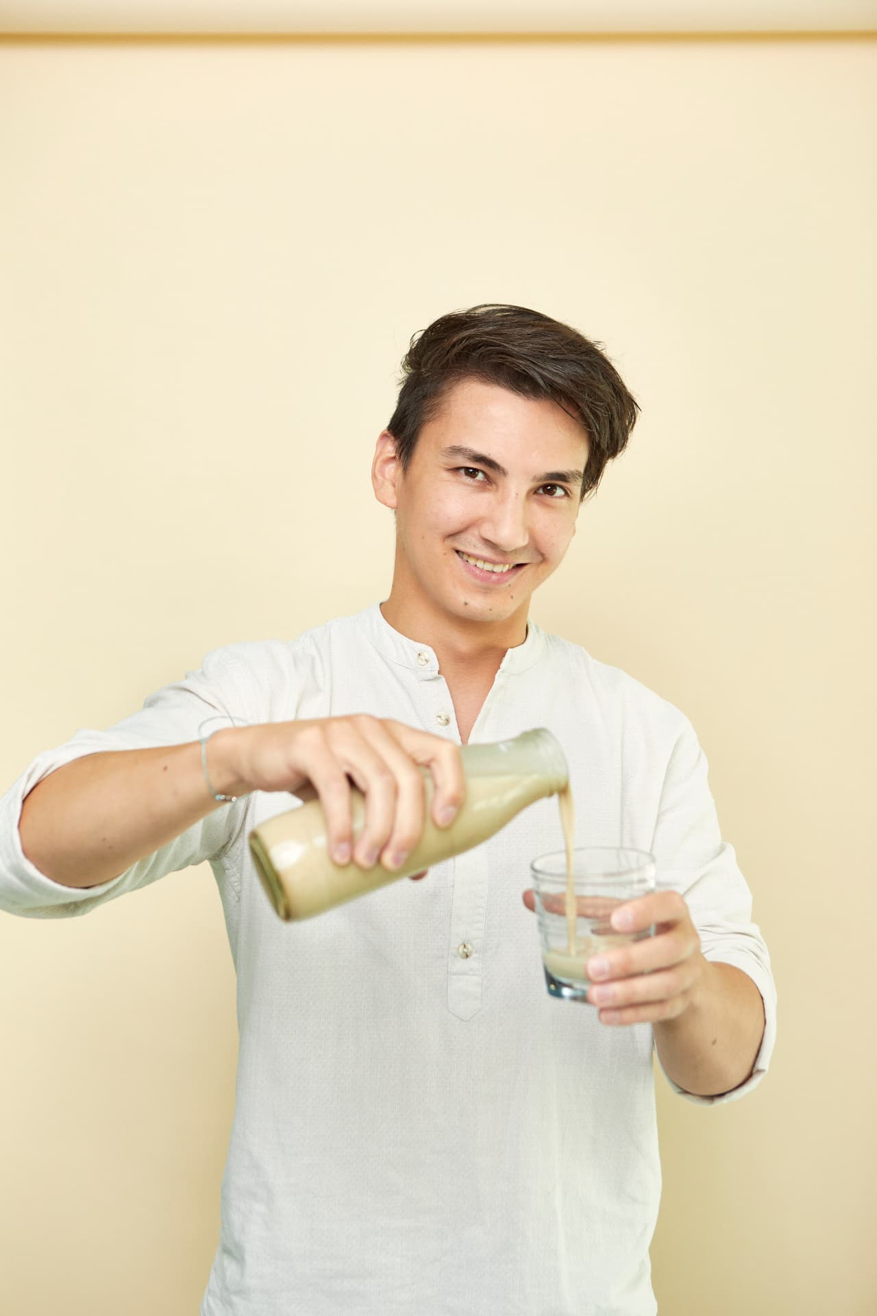 Elias Grau schenkt Haferdrink in ein Glas ein, cremefarbenes Hemd mit Stehkragen vor cremefarbenem Hintergrund