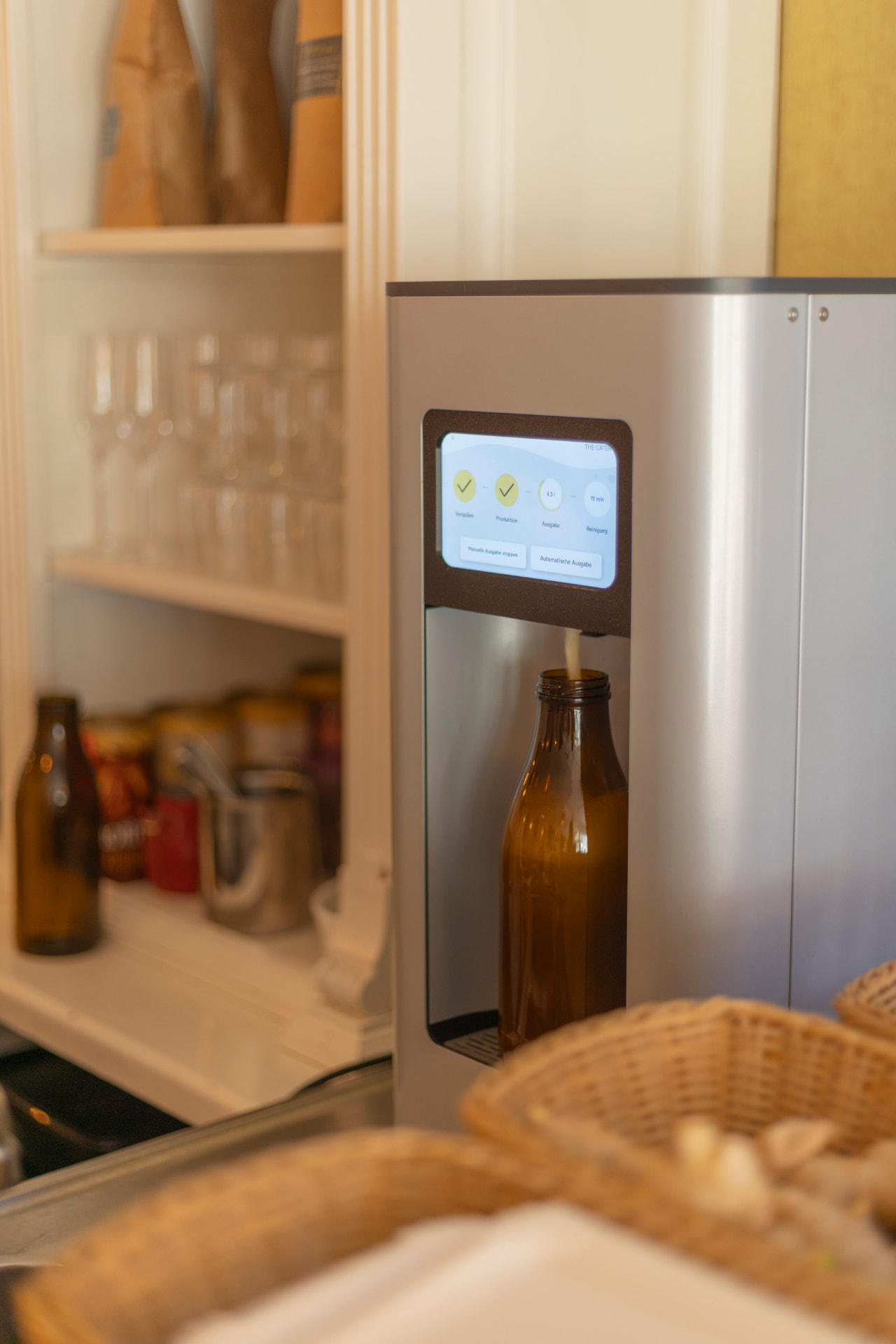 Die The Oater Maschine mit Touchscreen füllt frischen Haferdrink in eine braune Glasflasche, im Vordergrund unscharfe Weidenkörbe