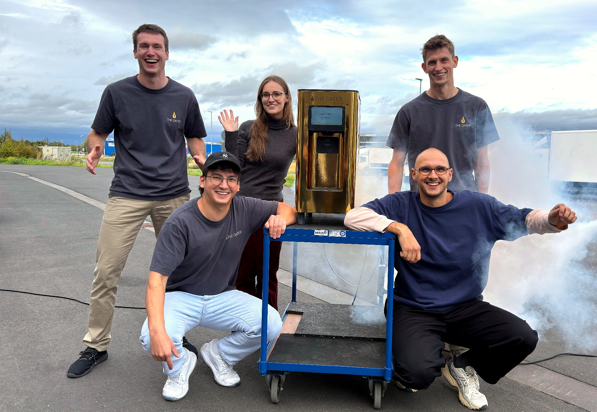 Fünf Teammitglieder posieren lachend mit der goldenen The Oater Maschine auf einem Transportroller