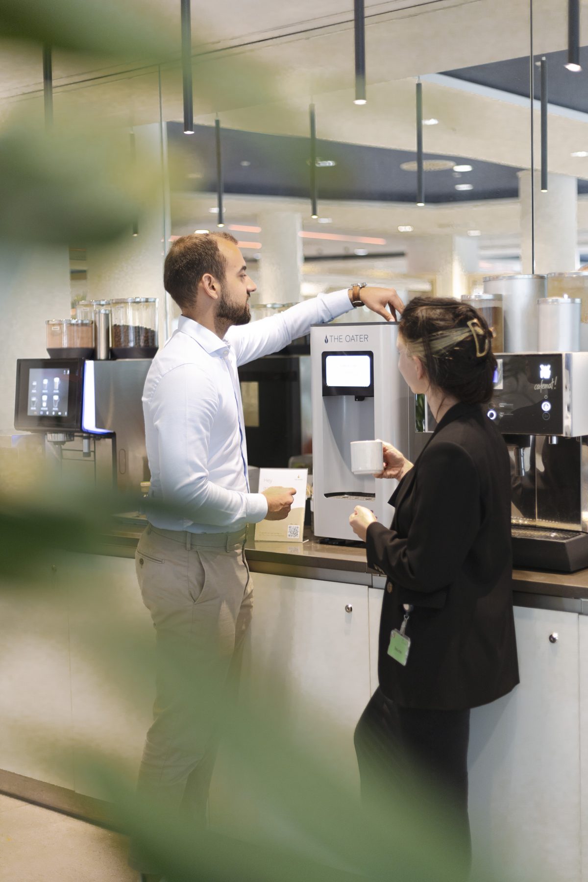 Zwei Personen mit Kaffeetassen stehen neben der The Oater Haferdrinkmaschine an einer Kaffeebar