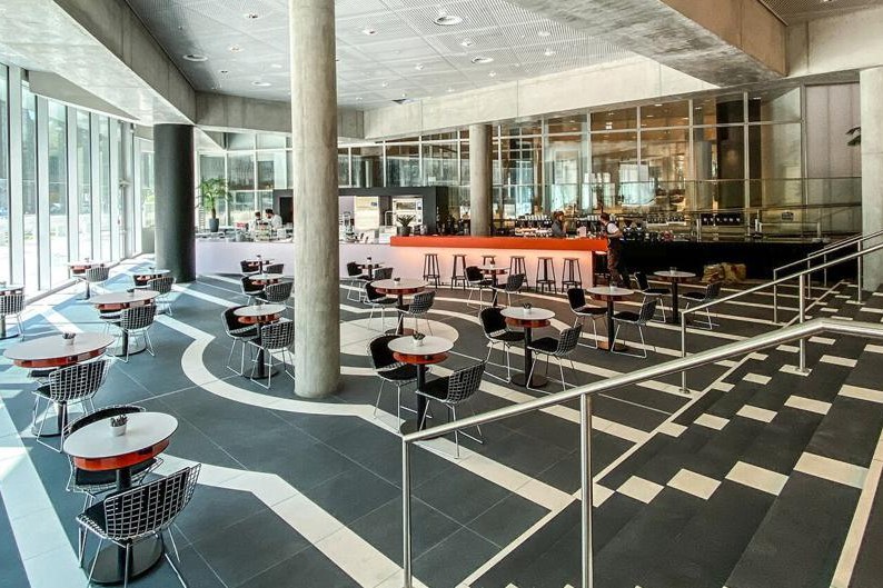 Großes helles Foyer mit roter Café-Theke, runden Tischen mit Drahtstühlen und grafischem Bodenmuster