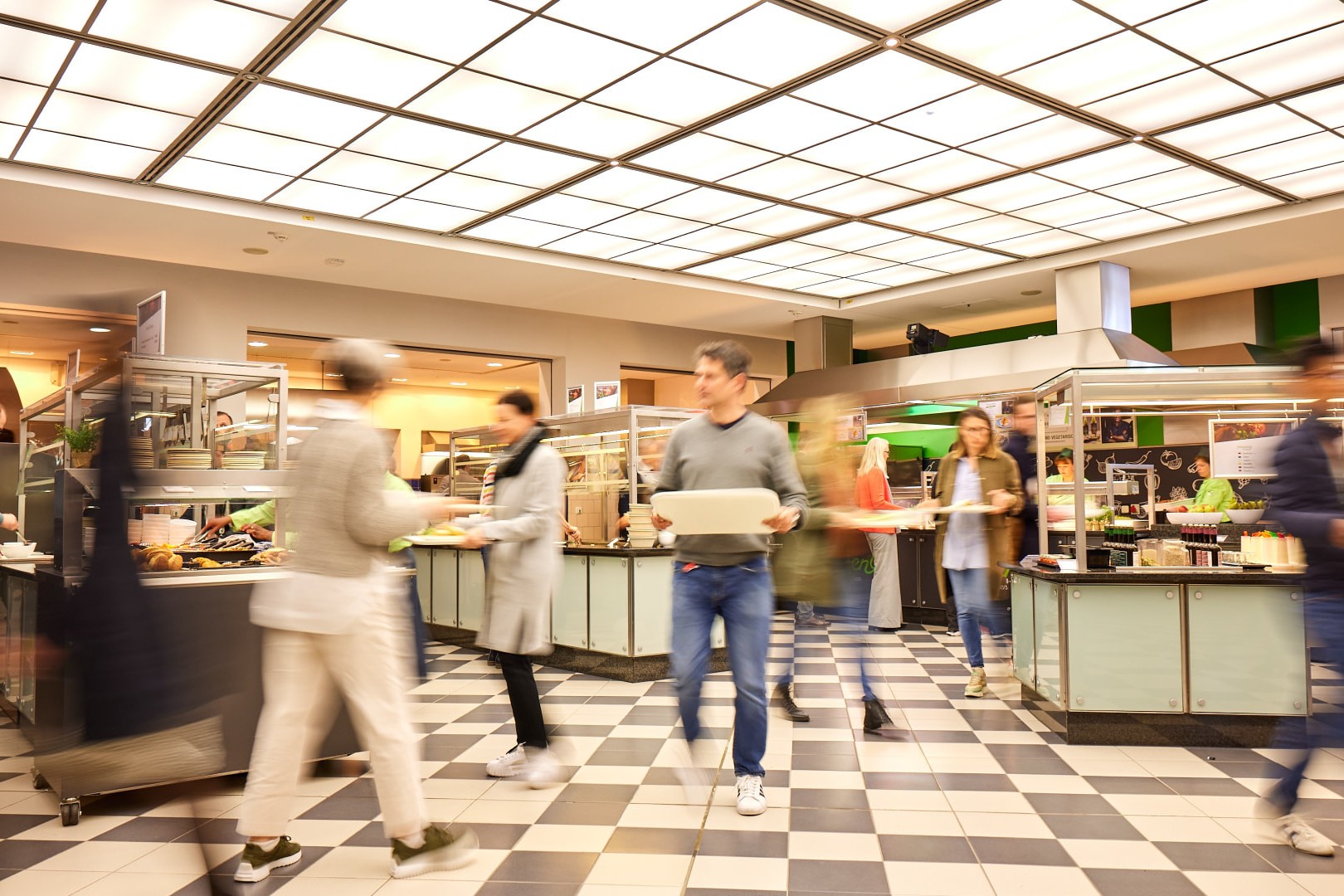 Helle Kantine mit Buffet- und Ausgabetheken unter einer großflächigen Raster-Lichtdecke, schwarz-weiß kariertem Boden und Gästen mit Tabletts