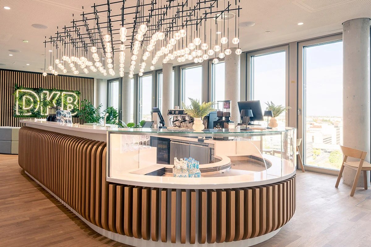 Café-Lounge der DKB mit geschwungenem Holztresen, Glasvitrine und Neon-Schriftzug DKB vor einer Mooswand