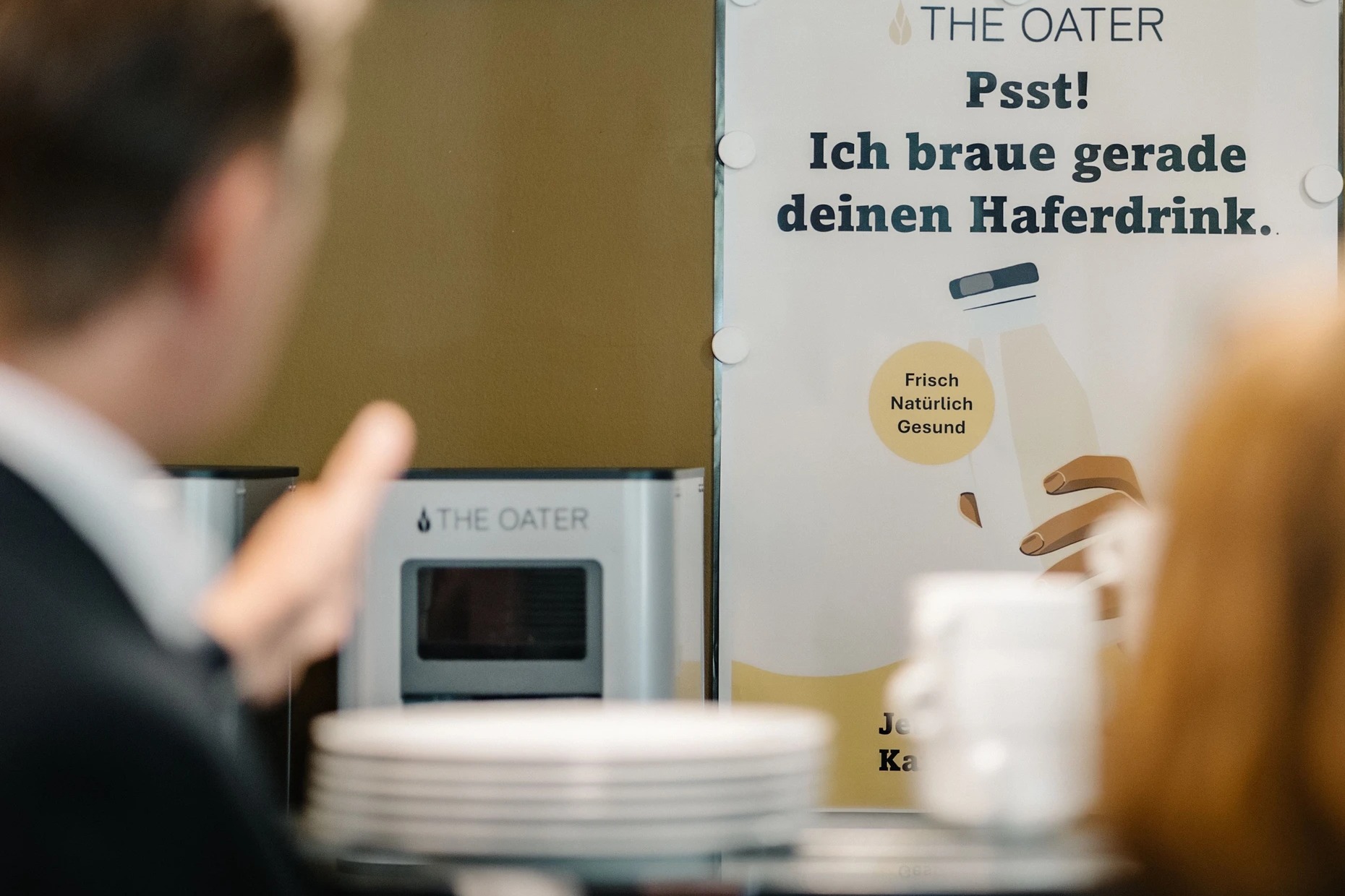 The-Oater-Maschine an einer olivgrünen Wand neben einem weißen Plakat mit der Aufschrift Psst! Ich braue gerade deinen Haferdrink, davor Geschirr und Tassen