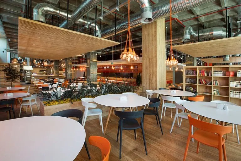 Helle Kantine mit runden weißen Tischen, orangen und grauen Schalenstühlen, hängenden Glühbirnenbündeln, Holzlamellen-Paneelen und einem Bücherregal