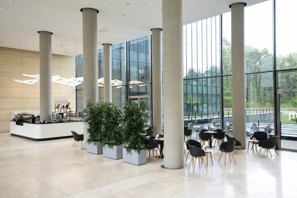 Hohes Foyer mit geschwungener Café-Theke, wellenförmiger Lichtinstallation, grauen Pflanzkübeln und Sitzgruppen vor raumhoher Glasfront