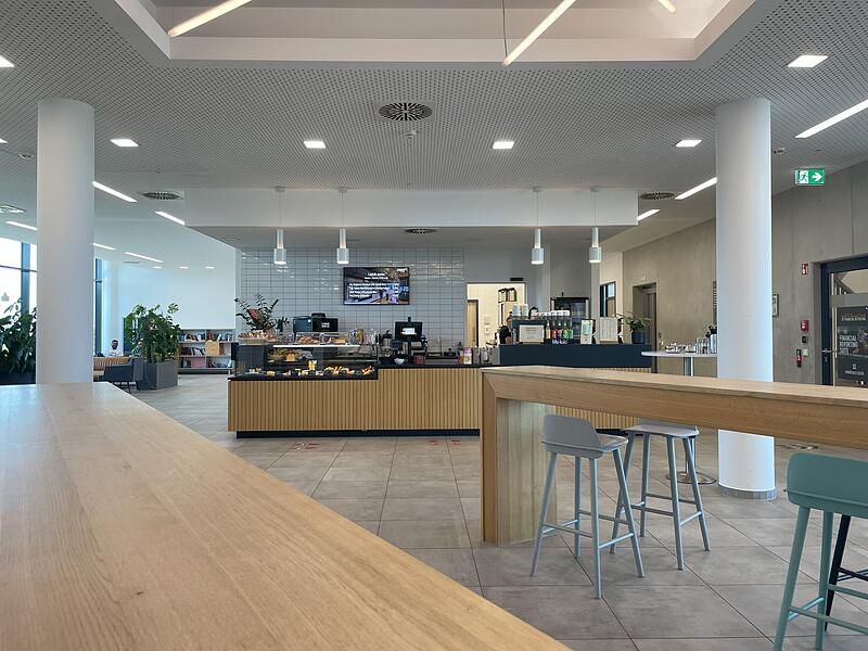 Heller Bistrobereich mit langem Holztresen, Barhockern und einer Snackvitrine vor weiß gefliester Wand