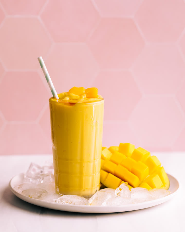 Vegan Mango Lassi im hohen Glas mit Mangowürfeln und Trinkhalm auf einem Teller mit Eis